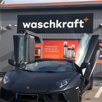 Schwarzer Sportwagen mit geöffneten Flügeltüren vor einer Waschkraft-Filiale, spezialisiert auf professionelle Fahrzeugpflege.