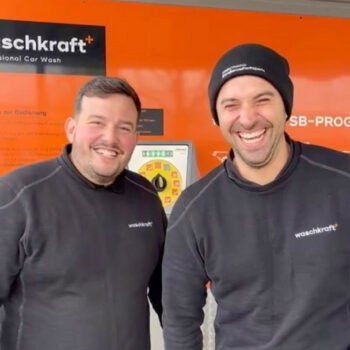 Zwei lachende Männer in waschkraft Kleidung vor einer orangefarbenen Wand mit einem Bedienfeld.