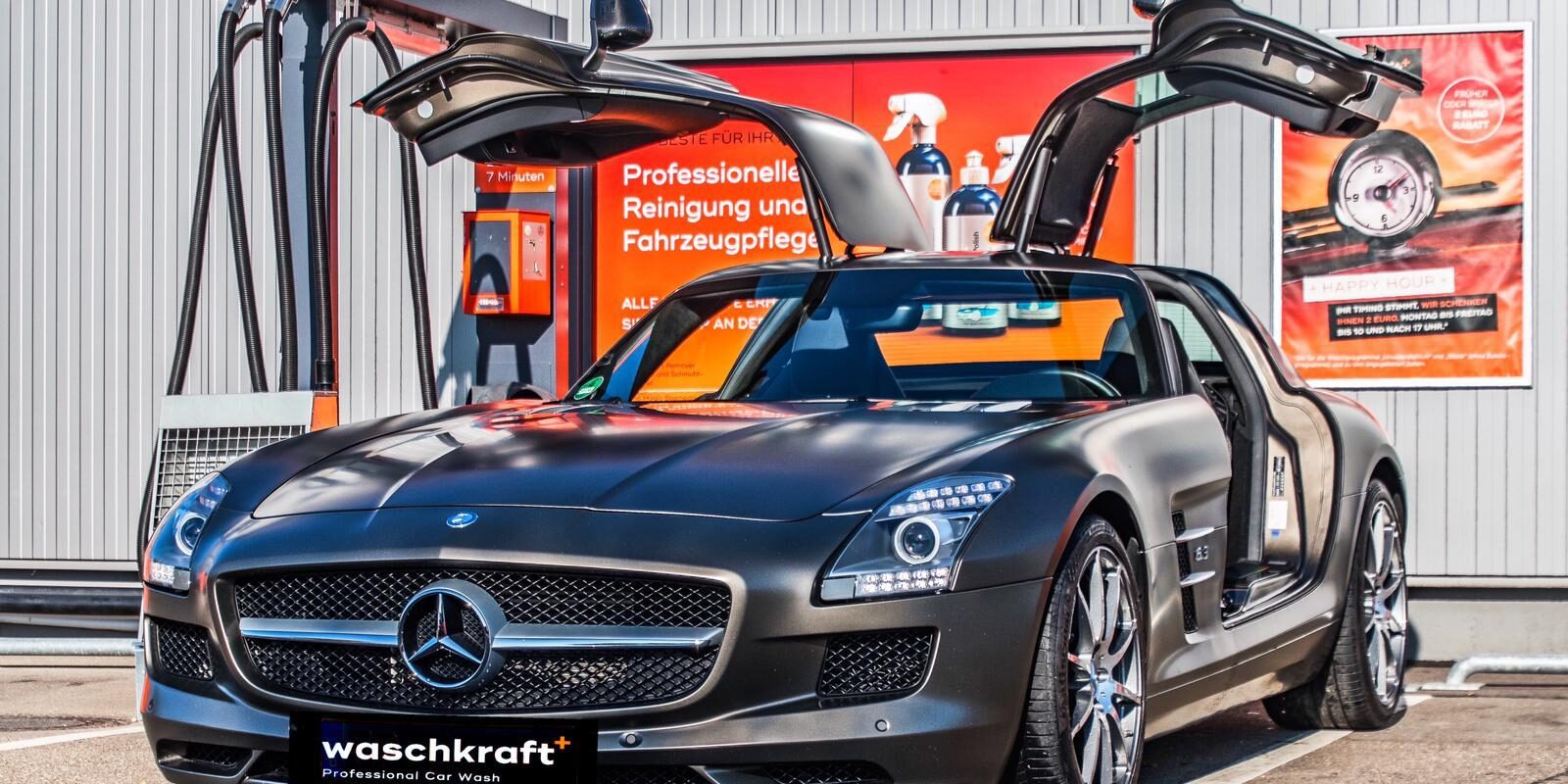Ein silbergrauer Sportwagen mit geöffneten Flügeltüren steht vor einer professionellen Autowaschanlage namens waschkraft.